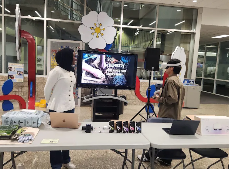 Tables with laptops on them and two people standing behind, one wearing a VR headset.