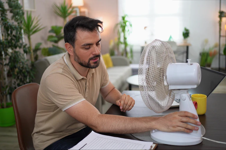 white man in beige T-shirt sits at office desk with fan