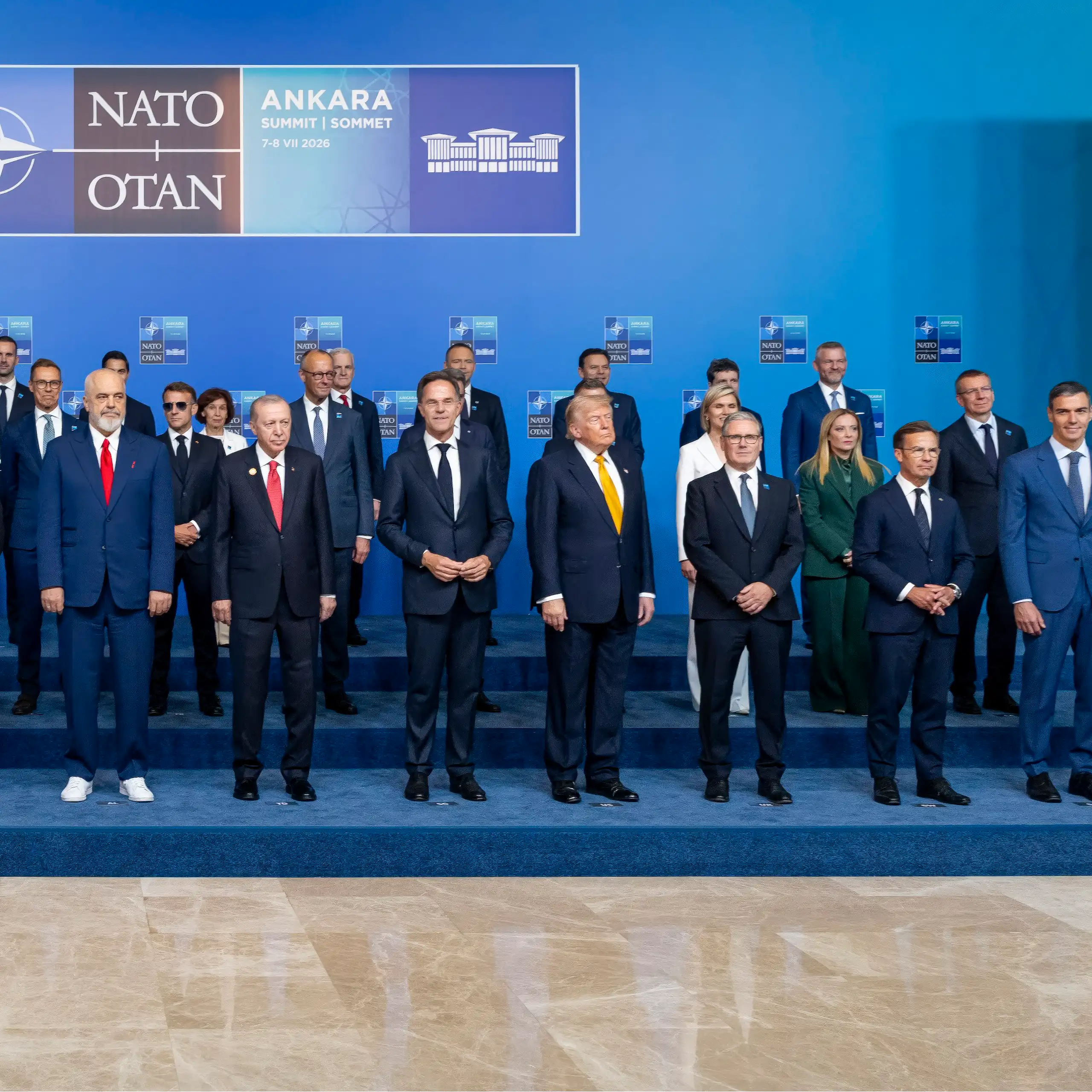 Multiple heads of state pose for a photo in front of a background that features the Nato logo