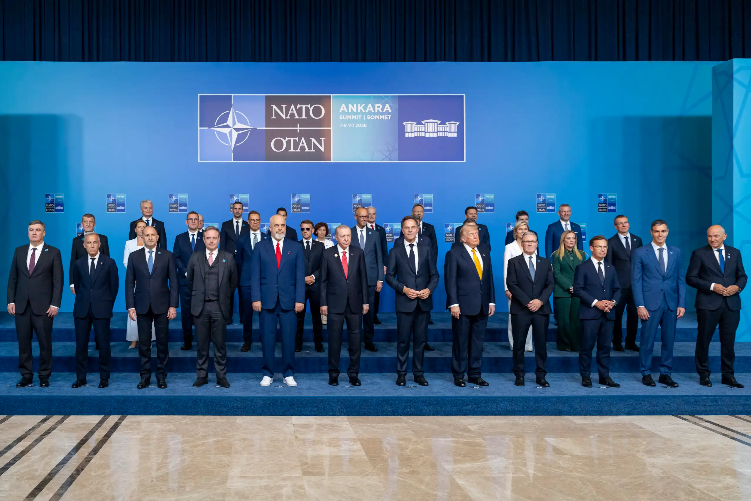 Multiple heads of state pose for a photo in front of a background that features the Nato logo