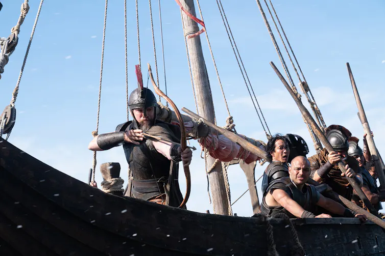 Un hombre apunta con un arco desde la cubierta de un barco hacia algo fuera de campo.