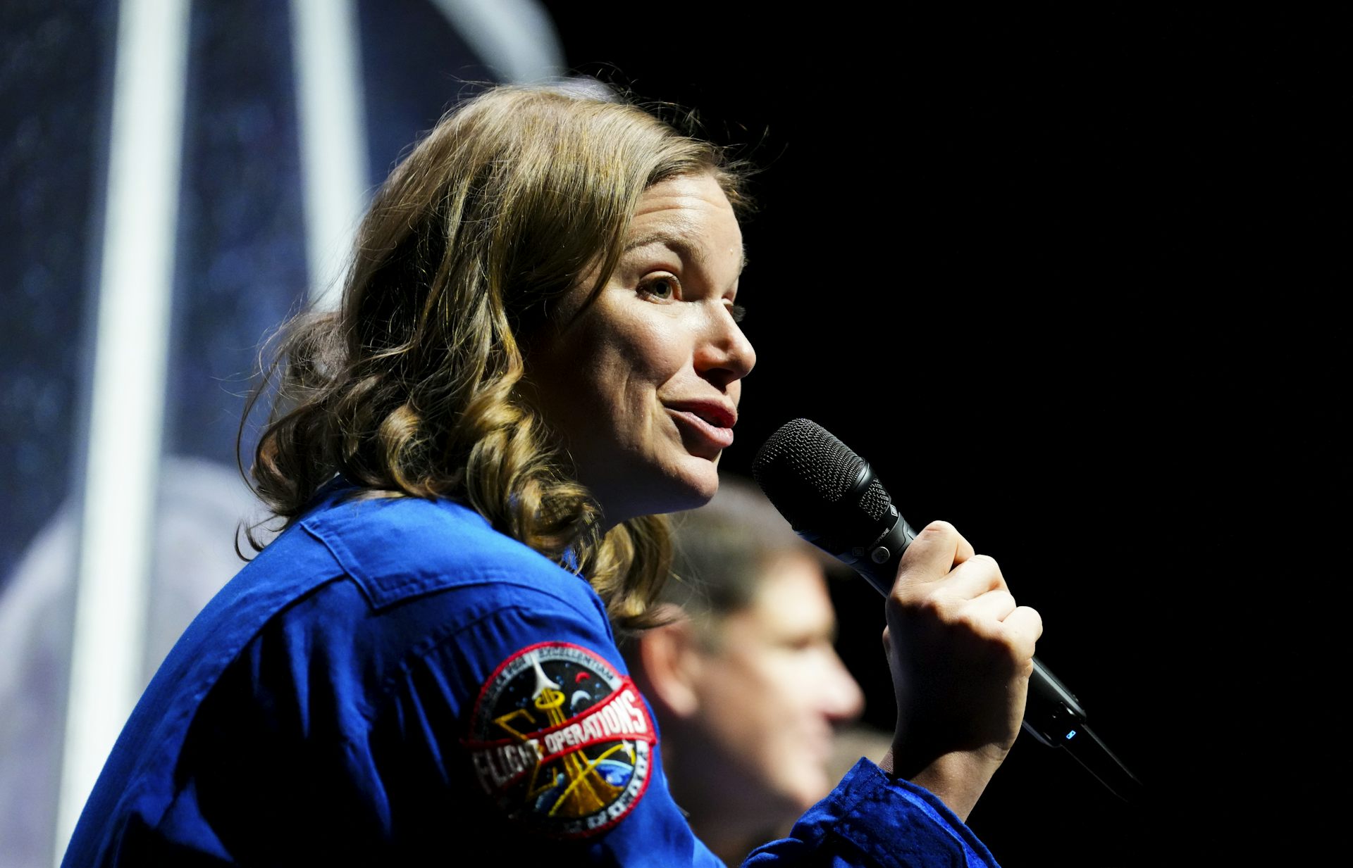 A woman with wavy dark blonde hair speaks into a microphone.