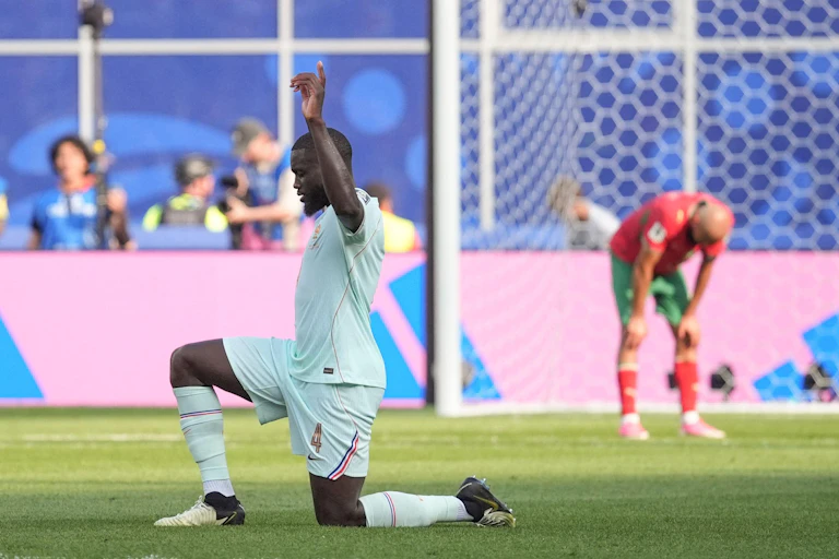 Dayot Upamecano faces fans down on one knee with a hand in the air as a goalkeeper bends over looking exhausted in the background