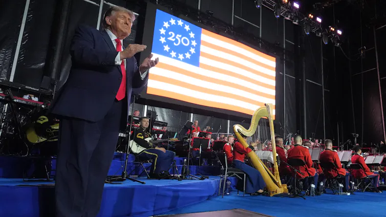 A man in a suit and red tie claps on stage with an American flag behind him.