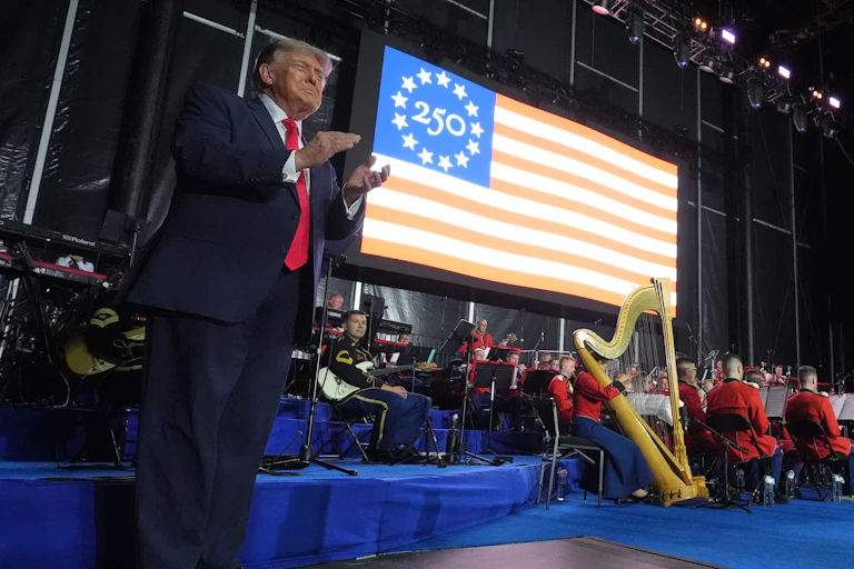 A man in a suit and red tie claps on stage with an American flag behind him.