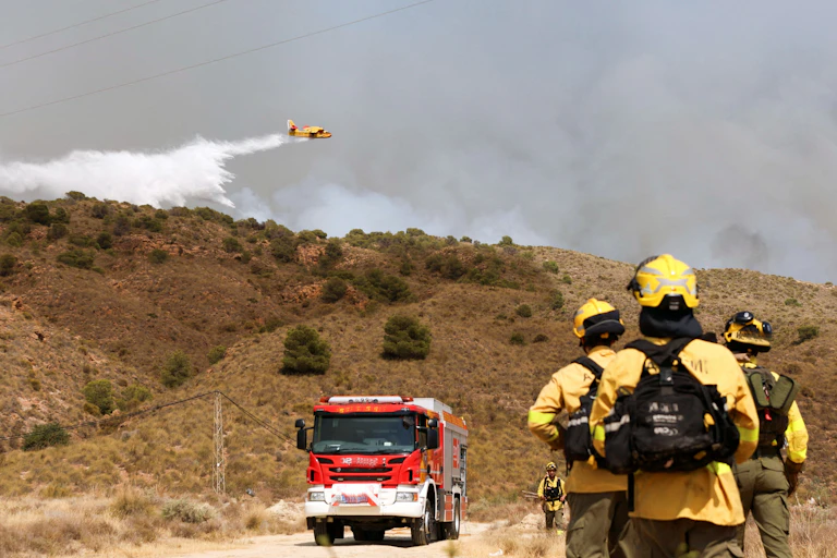 Tres bomberos de espaldas frente a un camión de bomberos observan un hidroavión echar agua sobre una zona detrás de una montaña de donde sale humo