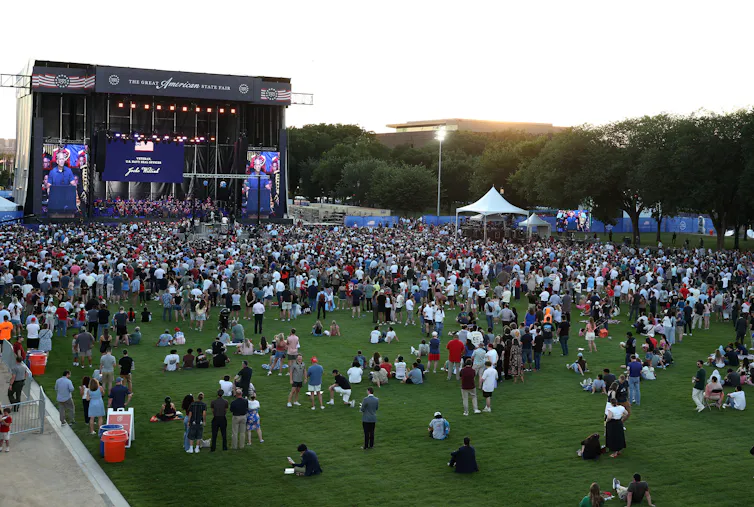 A crowd stands on a grass field in front of a stage.