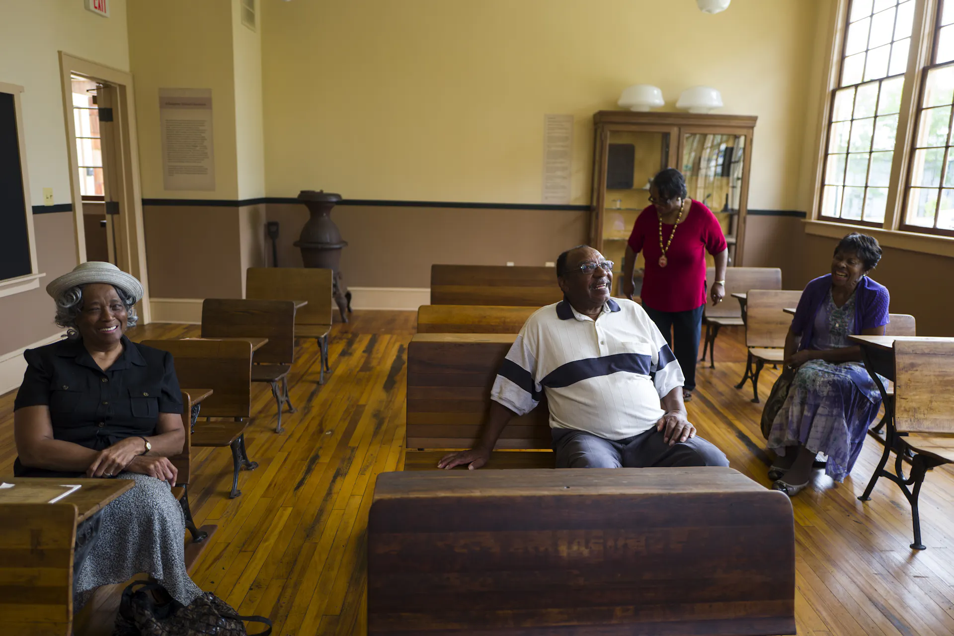 A group of older Black people gather in a restored old-fashioned schoolhouse.