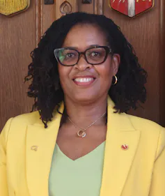 Una mujer negra sonriente con una chaqueta amarilla y una camisa verde pálido.