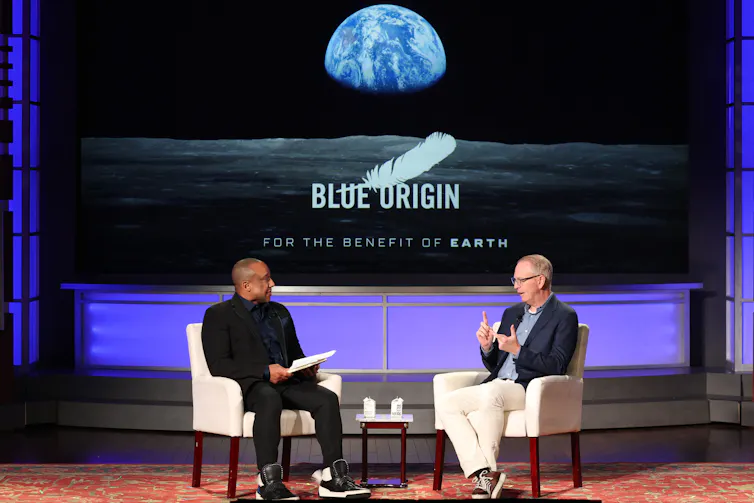 Two men sitting in chairs on a stage, with a screen behind them displaying the Blue Origin logo with a photo of the Earth from the Moon.