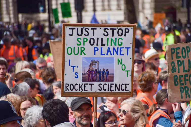 Protestors march through a London street. One holds a sign reading 'Just Stop Spoiling our Planet, it's not Rocket Science'