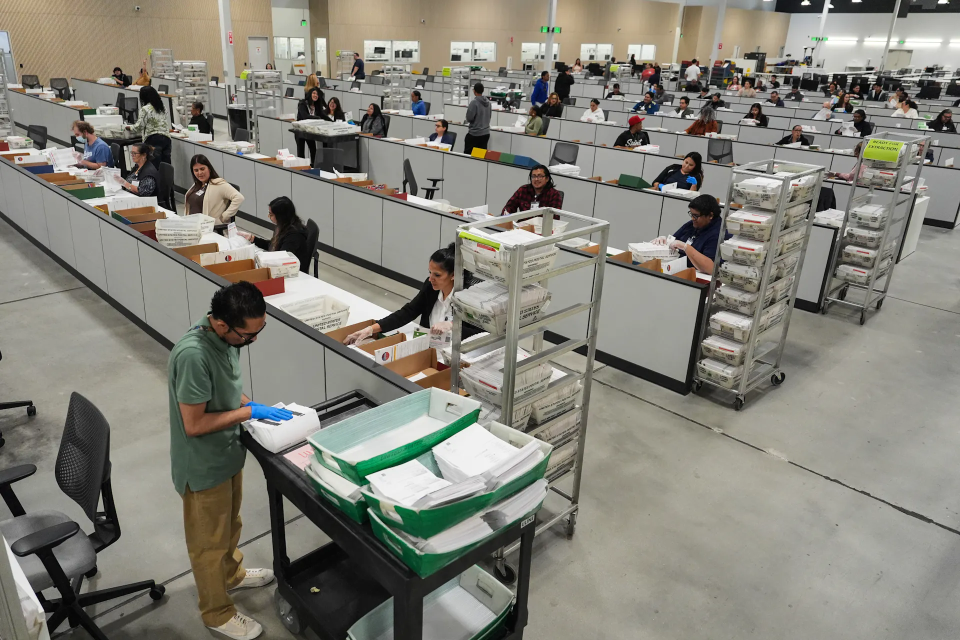 Rows of long desks fill a large room where workers are sitting and inspecting ballots. One worker stands in the foreground.