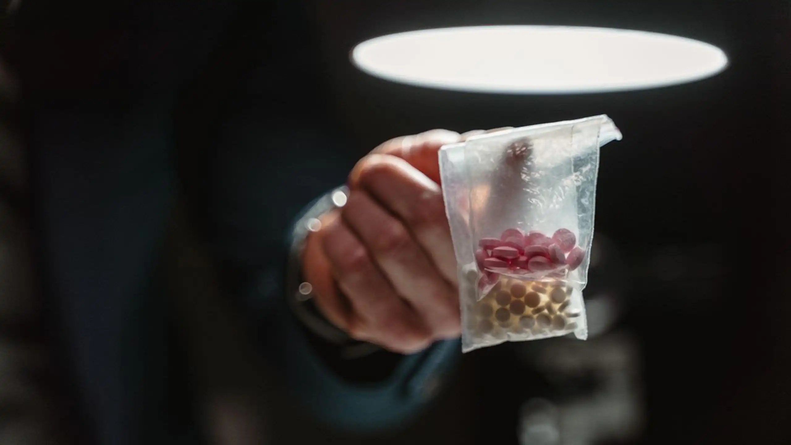 Finger holding up bag of pills, in darkened room with ceiling light