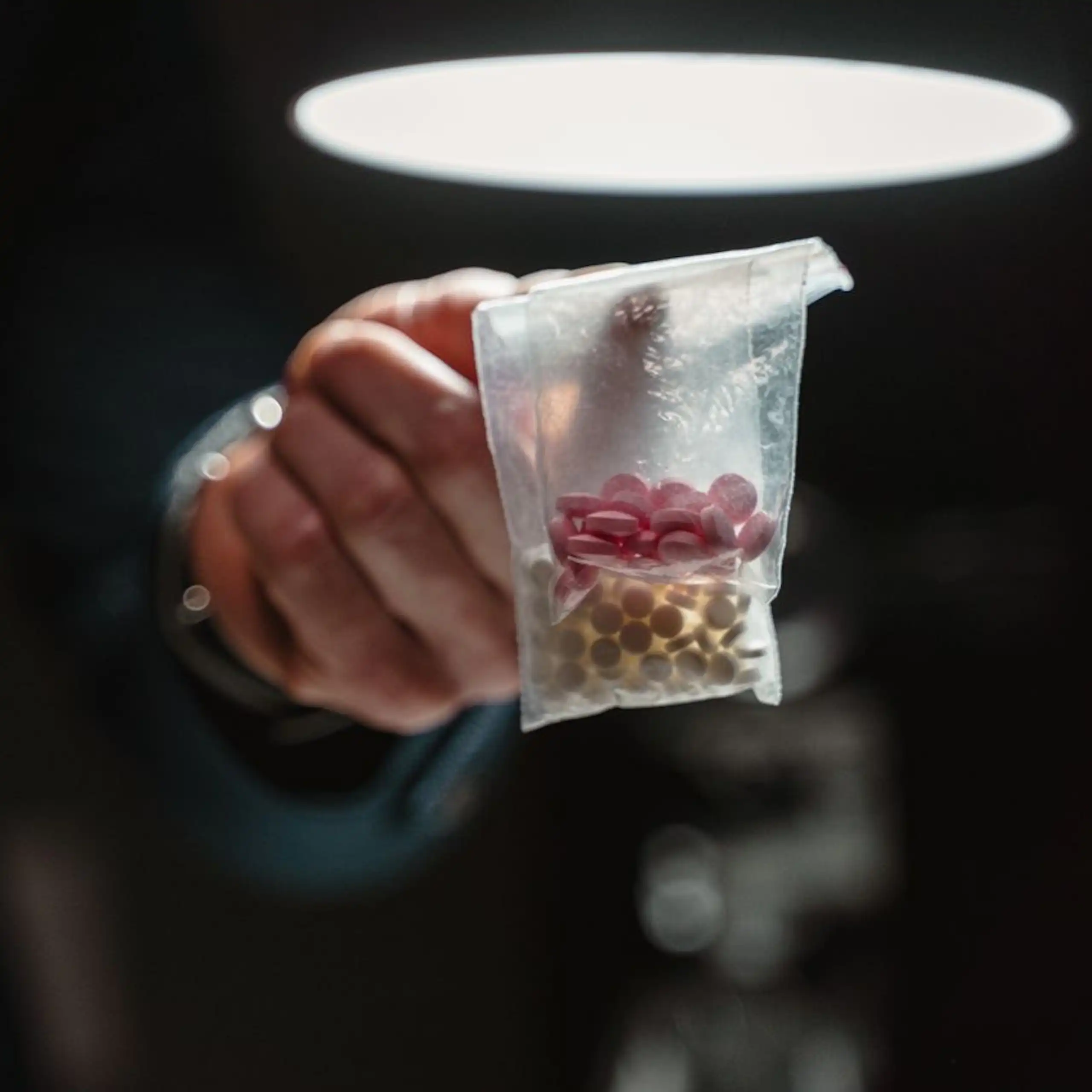 Finger holding up bag of pills, in darkened room with ceiling light