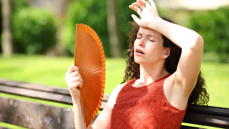 Una mujer pasando calor sentada en un banco con un abanico y la otra mano en la frente