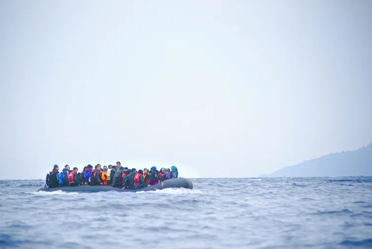 A colour photograph of people on an inflatable dinghy on a grey sea with a sliver of land in the background.