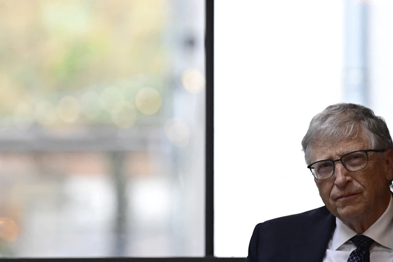 Bill Gates looks contemplative while sitting in front of a window.