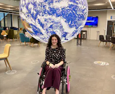 A brown haired young woman in a wheelchair with pink wheels and flowers smiles in front of a large illuminated globe suspended from the ceiling of Space Park Leicester, University of Leicester.