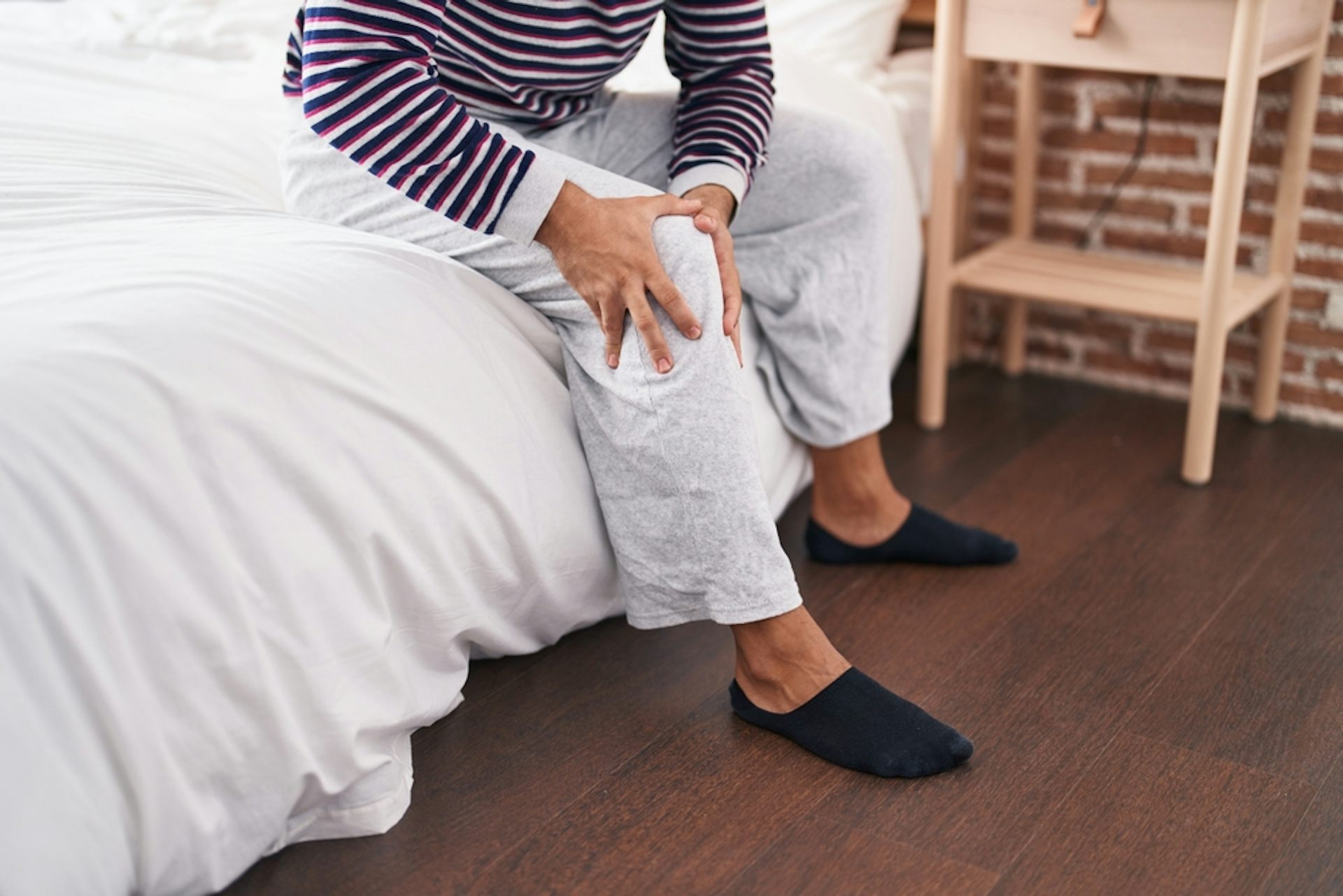 A person sitting on the edge of a bed, rubbing their knee.