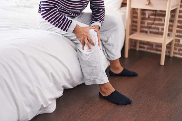 A person sitting on the edge of a bed, rubbing their knee.