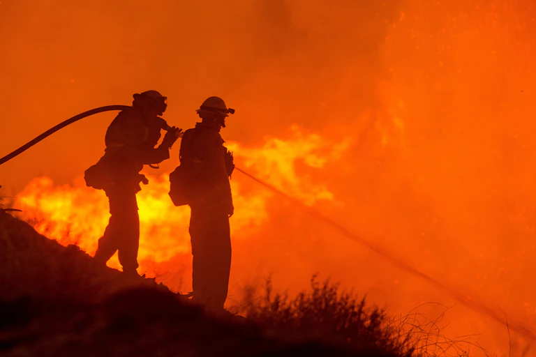 silhouettes of two firefighters with hose, spraying red flames on hill