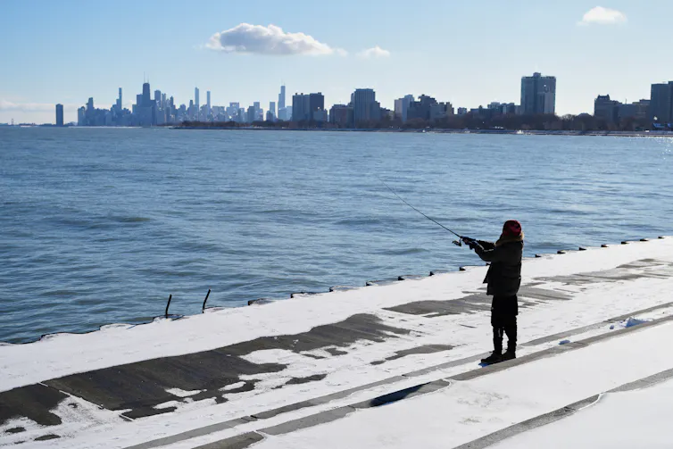 A person stands on a flat area and holds a fishing rod out over a body of water with a city skyline in the background.