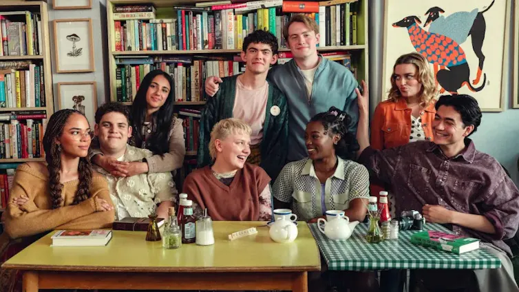 Cast of Heartstopper sitting together against a bookshelf and two tables with diner items