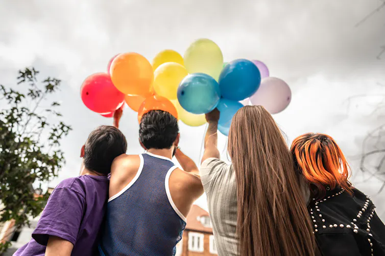 Group of four people from behind standing outside, holding an array of balloons in a rainbow arc