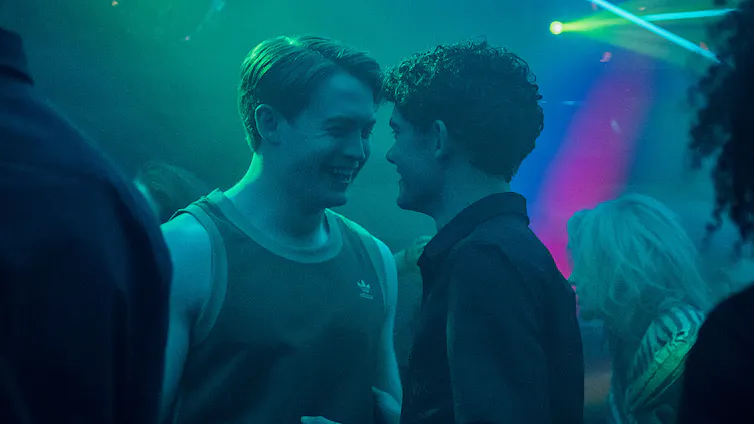Two boys smiling at each other in a dim room bathed in neon lights