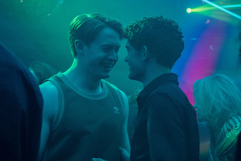 Two boys smiling at each other in a dim room bathed in neon lights