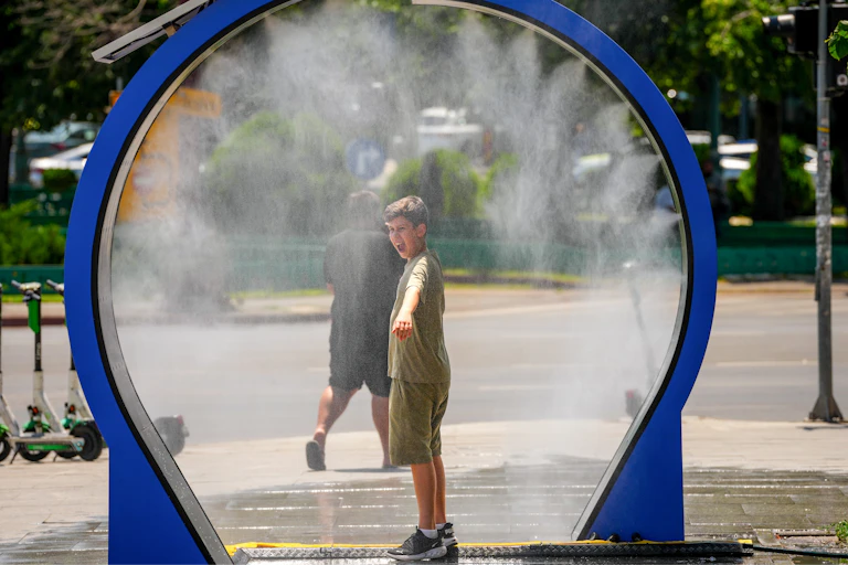 Un jeune garçon se fait asperger d'eau sous un brumisateur, installé dans un espace public près d'un trottoir.