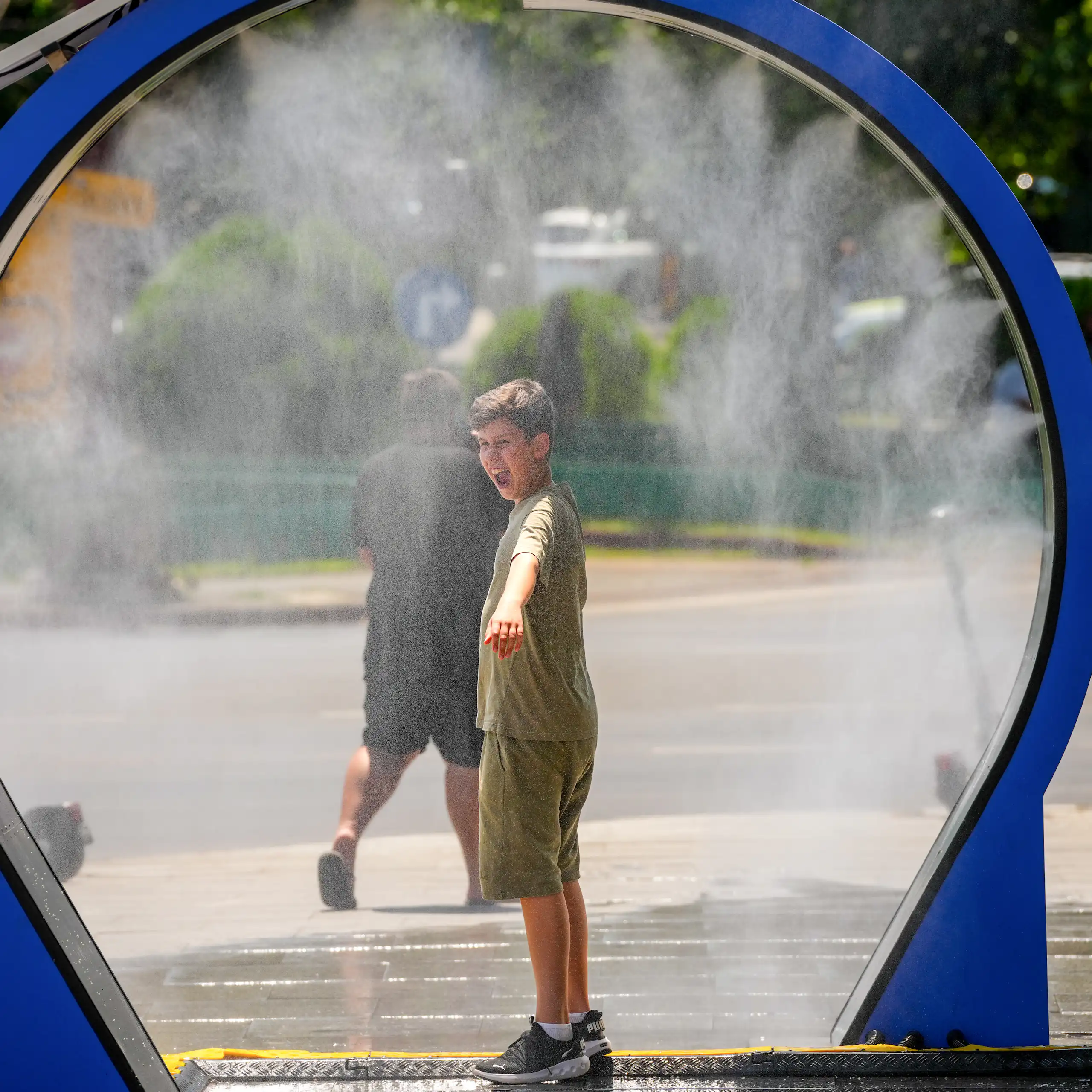 Un jeune garçon se fait asperger d'eau sous un brumisateur, installé dans un espace public près d'un trottoir.