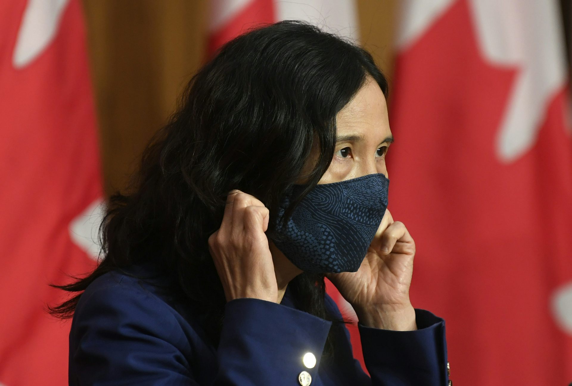 La Dre Theresa Tam, est photographiée en gros plan, portant un masque, avec un drapeau du Canada à l’arrière-plan