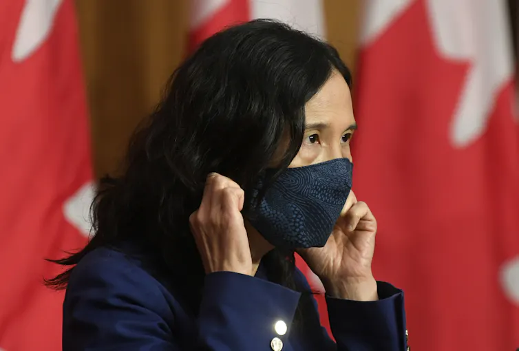 La Dre Theresa Tam, est photographiée en gros plan, portant un masque, avec un drapeau du Canada à l’arrière-plan