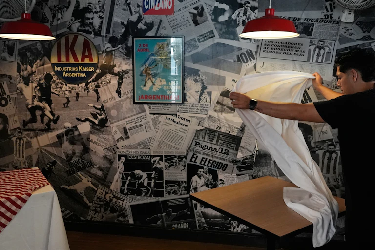 A waiter sets up tables at an Argentine restaurant decorated with sports newspaper clippings, including coverage of Diego Maradona during the 1986 World Cup.
