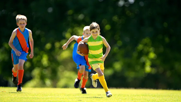 Three kids in jerseys play Australian rules footy on a field