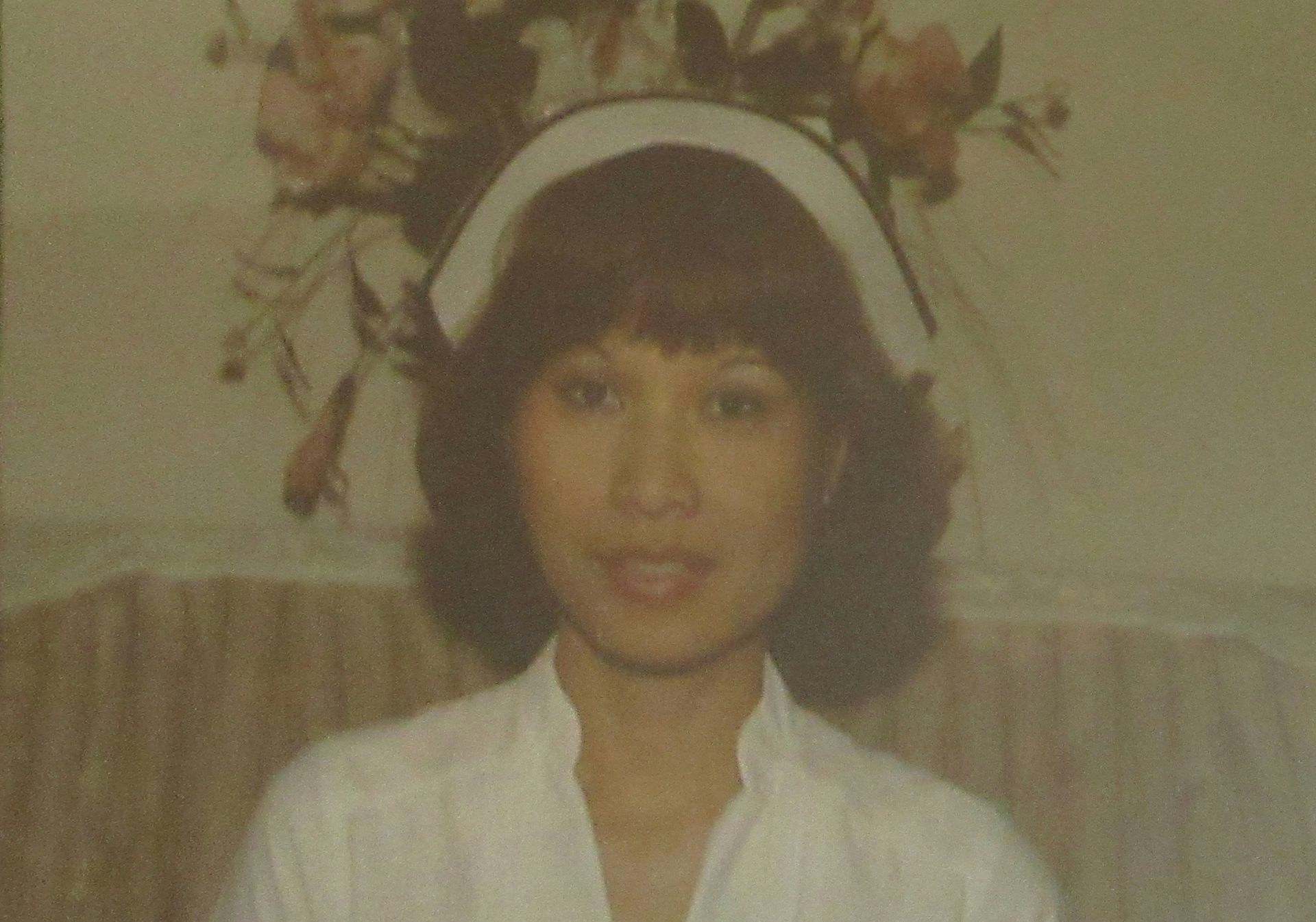 La foto muestra a una mujer de cabello oscuro con uniforme de enfermería.