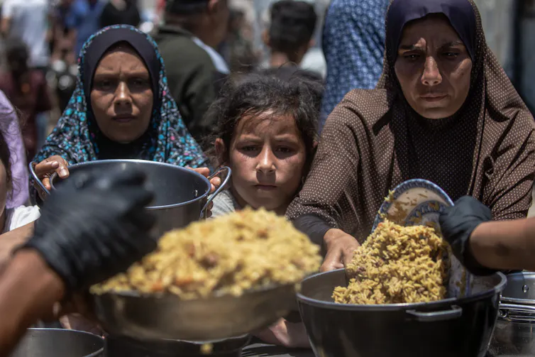 Two Muslim women and a girl hold out pots to receive food aid