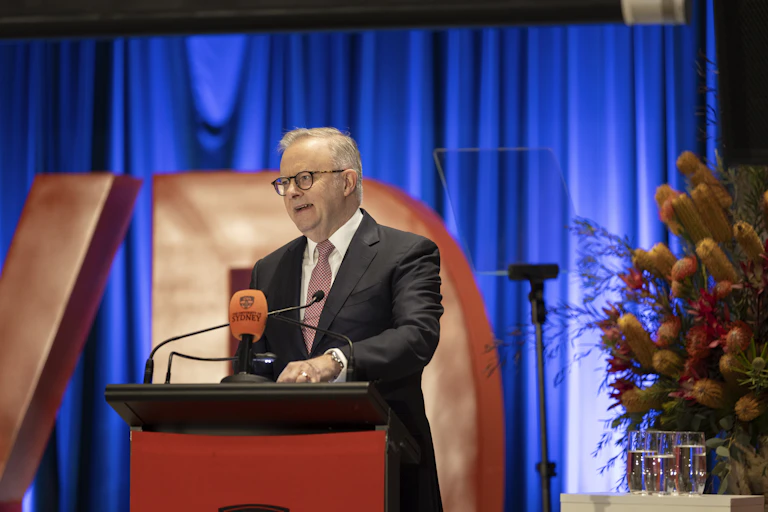 A man in a black suit speaking at lecturn, with flowers in the background.