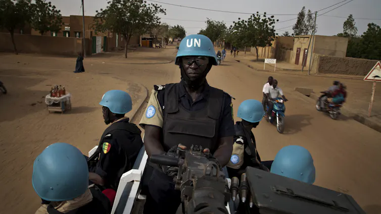 Five men wearing army fatigues and blue helmets with the letters UN in white, seated on the back of a vehicle
