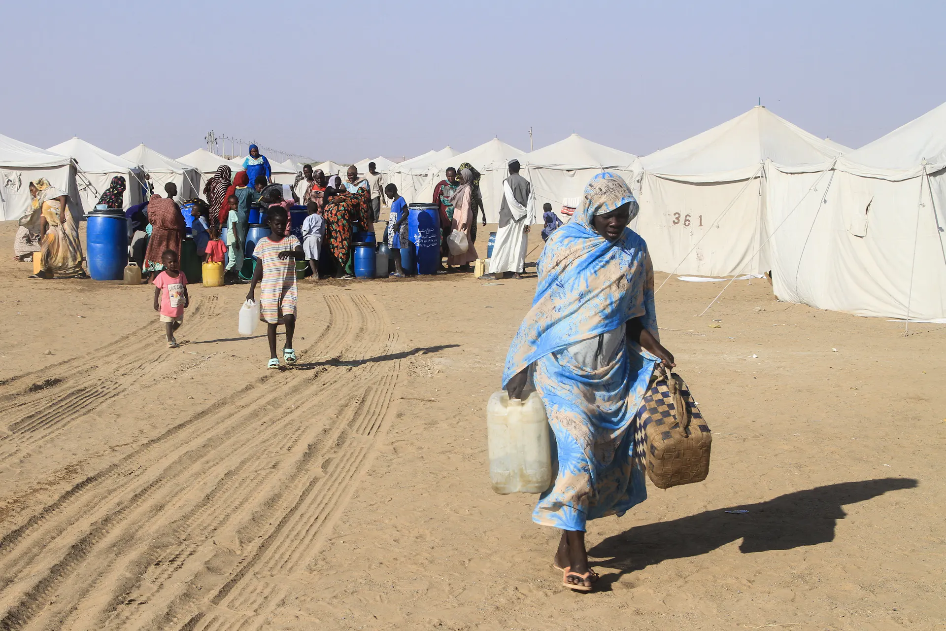 A woman carries cans of water in front of a row of tents.