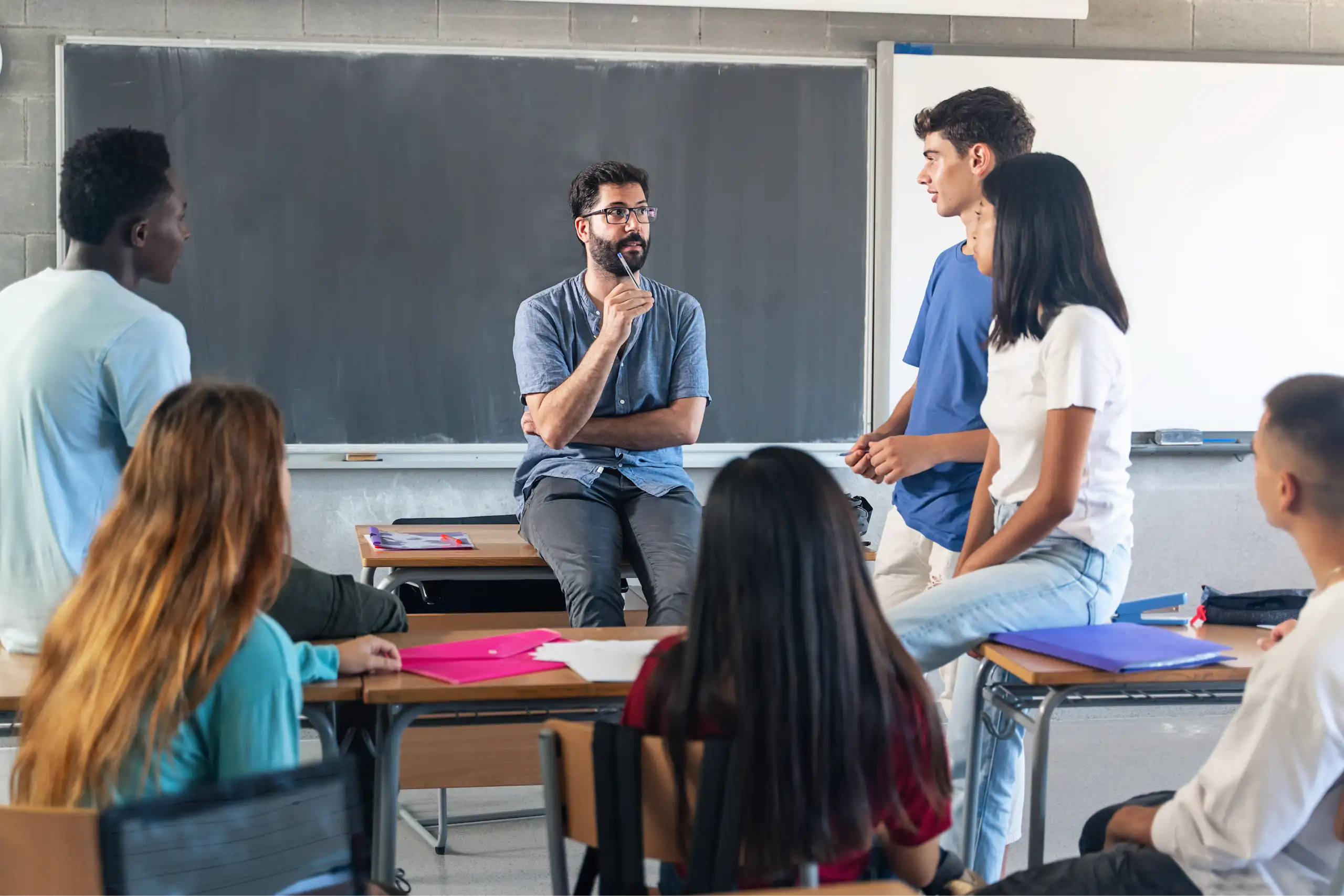 Un profesor explica en clase delante de la pizarra sentado en una mesa con varios alumnos de pie y sentados alrededor