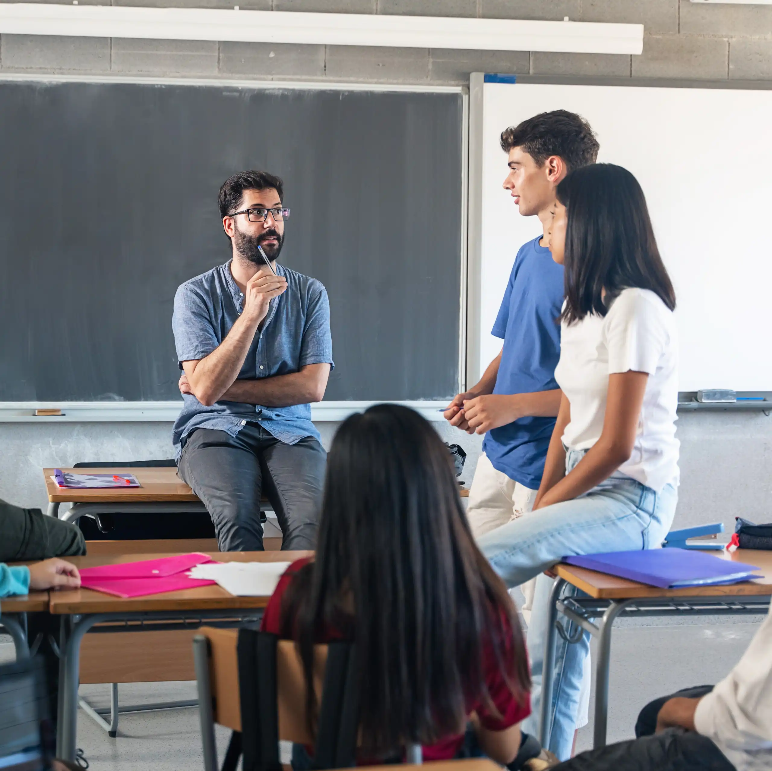 Un profesor explica en clase delante de la pizarra sentado en una mesa con varios alumnos de pie y sentados alrededor