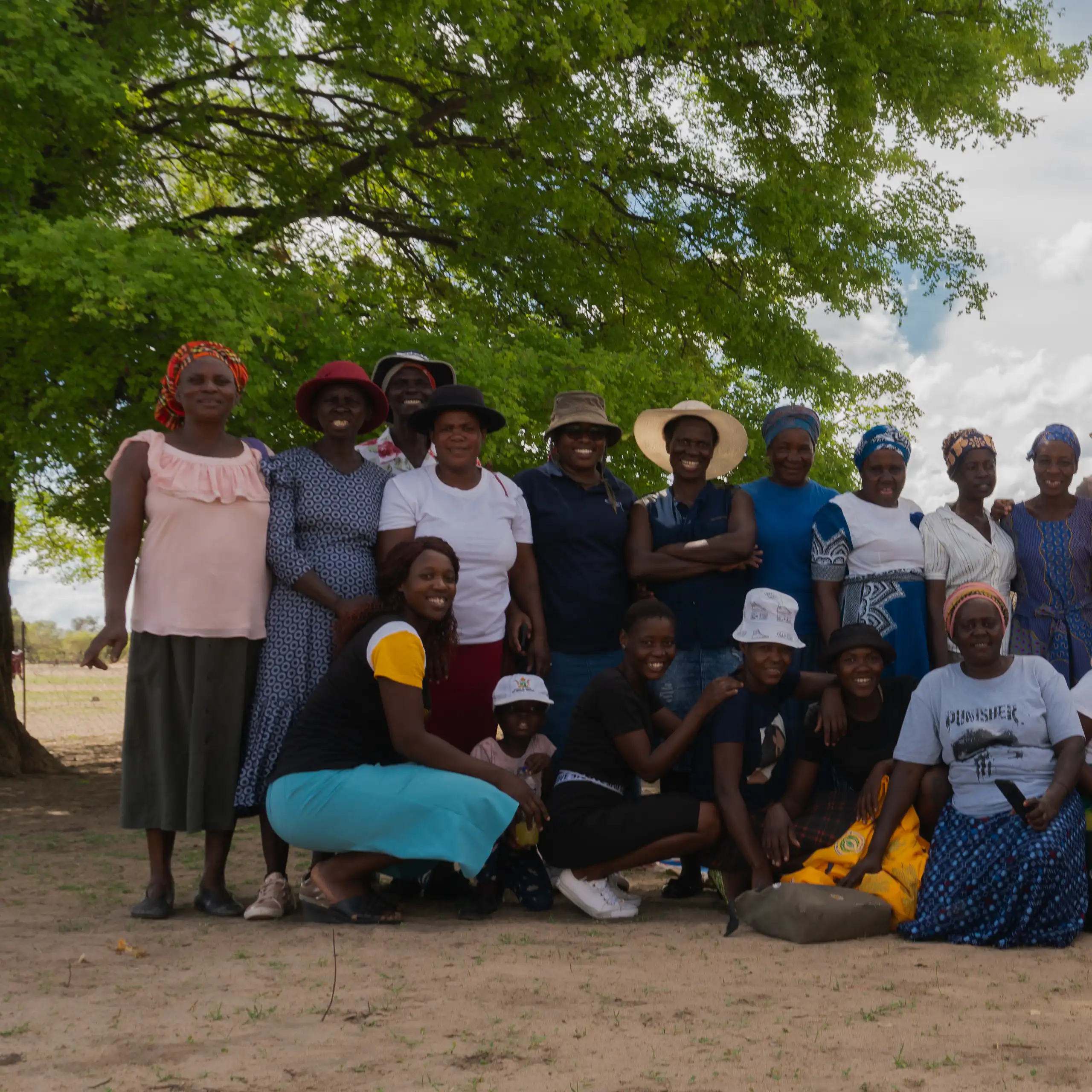 A group of about 25 women pose for a photo outside on dusty soil under a tree