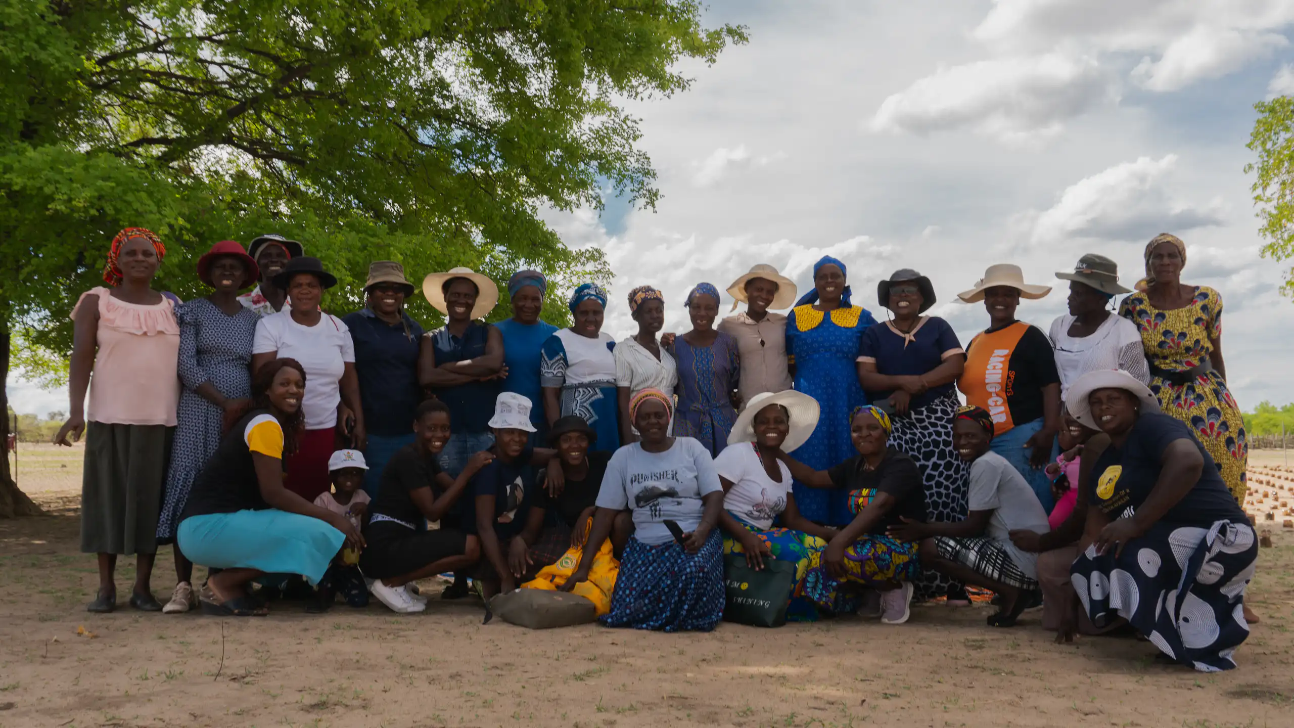 A group of about 25 women pose for a photo outside on dusty soil under a tree
