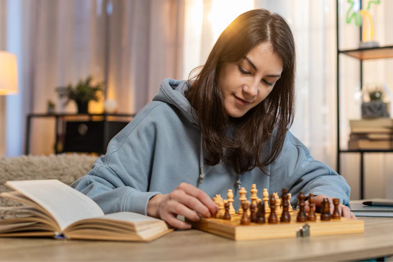 Una joven juega al ajedrez con un libro al lado en la mesa