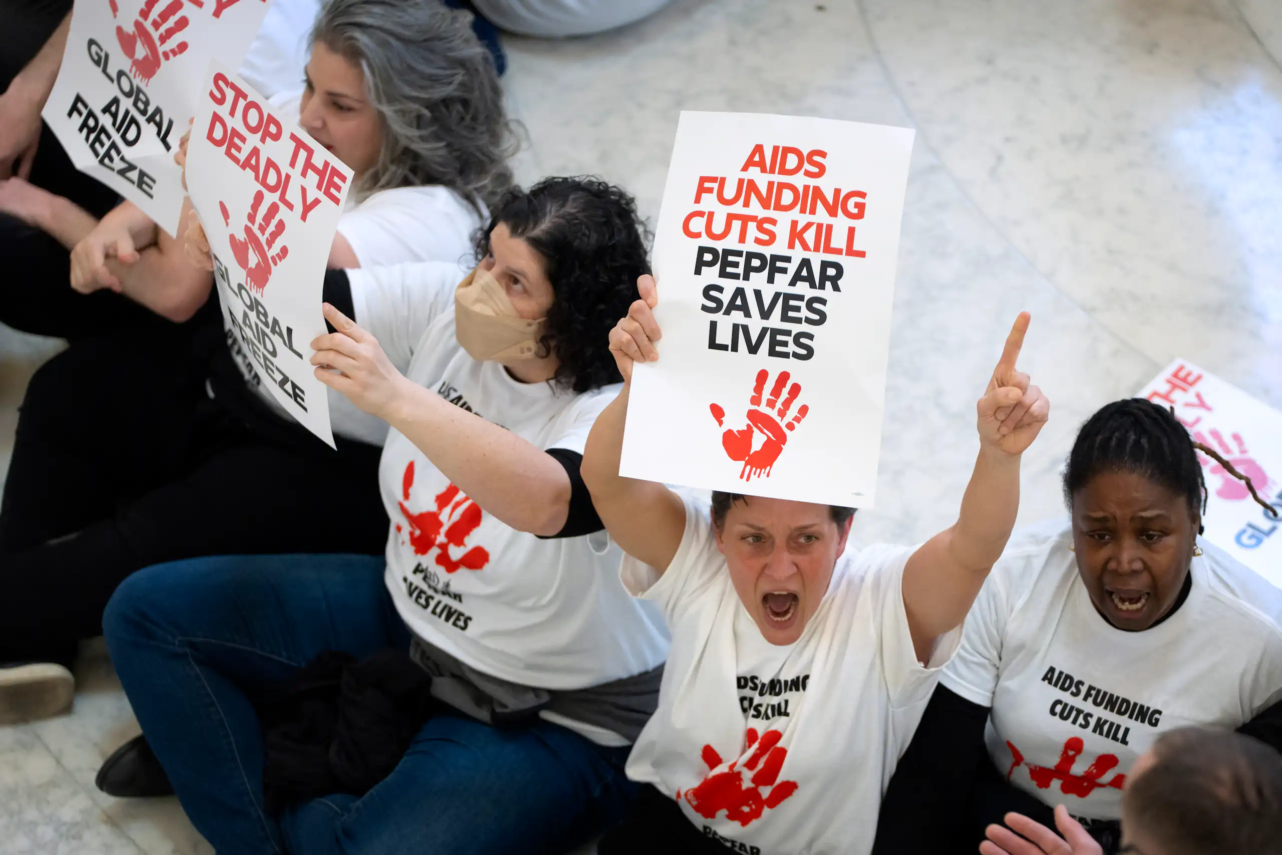 Demontrators sit on a stone floor holding signs decrying cuts to HIV/AIDS and foreign aid funding.