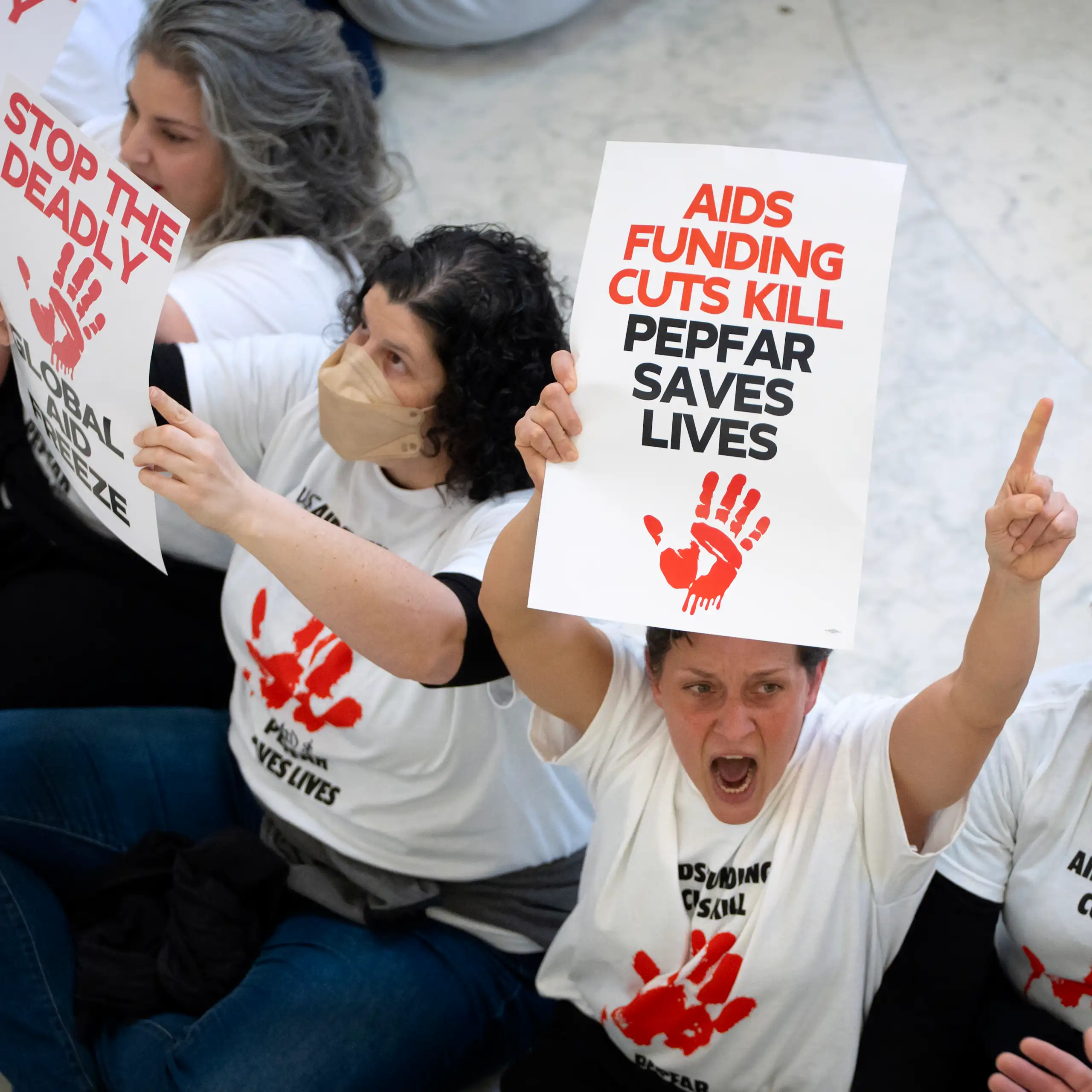 Demontrators sit on a stone floor holding signs decrying cuts to HIV/AIDS and foreign aid funding.