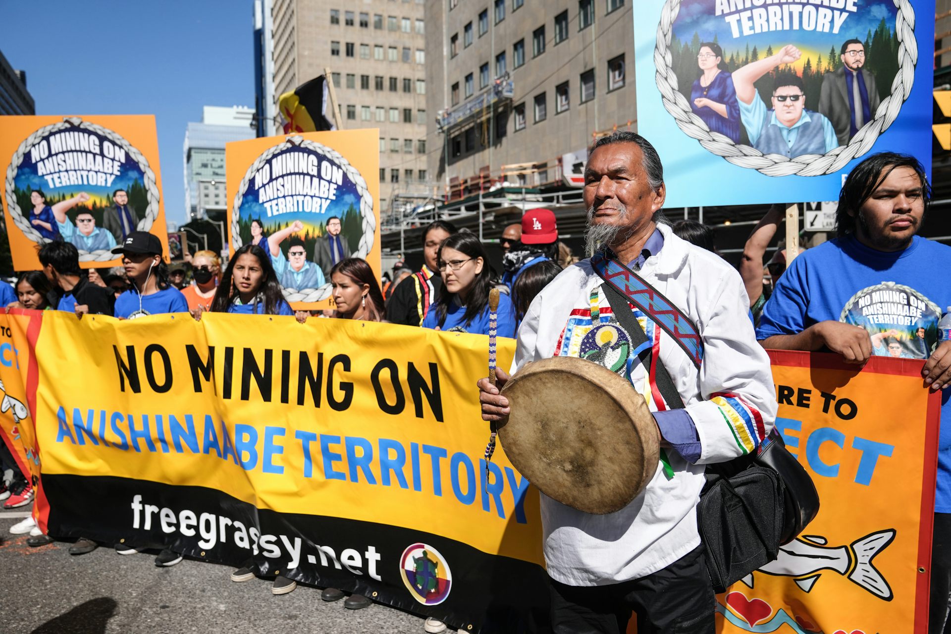 Un groupe d'autochtones brandit des bannières avec des slogans s'opposant aux mines, et l'un deux, à l'avant-plan, tient un tambour traditionnel.
