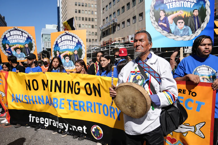 Un groupe d'autochtones brandit des bannières avec des slogans s'opposant aux mines, et l'un deux, à l'avant-plan, tient un tambour traditionnel.